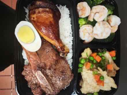 Three-compartment bento box with roast chicken leg, sliced beef over rice, shrimp with broccoli, and cauliflower with peas and carrots