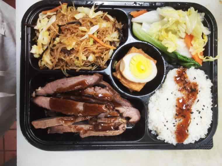 Bento box with stir-fried chow mein noodles, cabbage and green pepper salad, a halved boiled egg on braised tofu, sliced glazed roast meat and rice