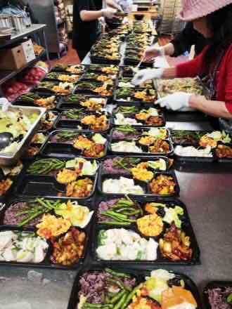 Kitchen counter lined with black compartment bento boxes holding purple rice, green beans, scrambled egg, steamed fish and diced chicken as staff fill them