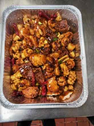 Catering tray of squid pieces and shrimp coated in a glossy dark red chili sauce with whole dried chilies and scallions