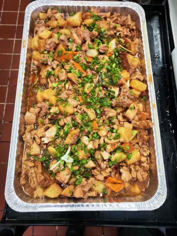 Large catering tray of braised chicken with potato and carrot in brown sauce, garnished with chopped scallion